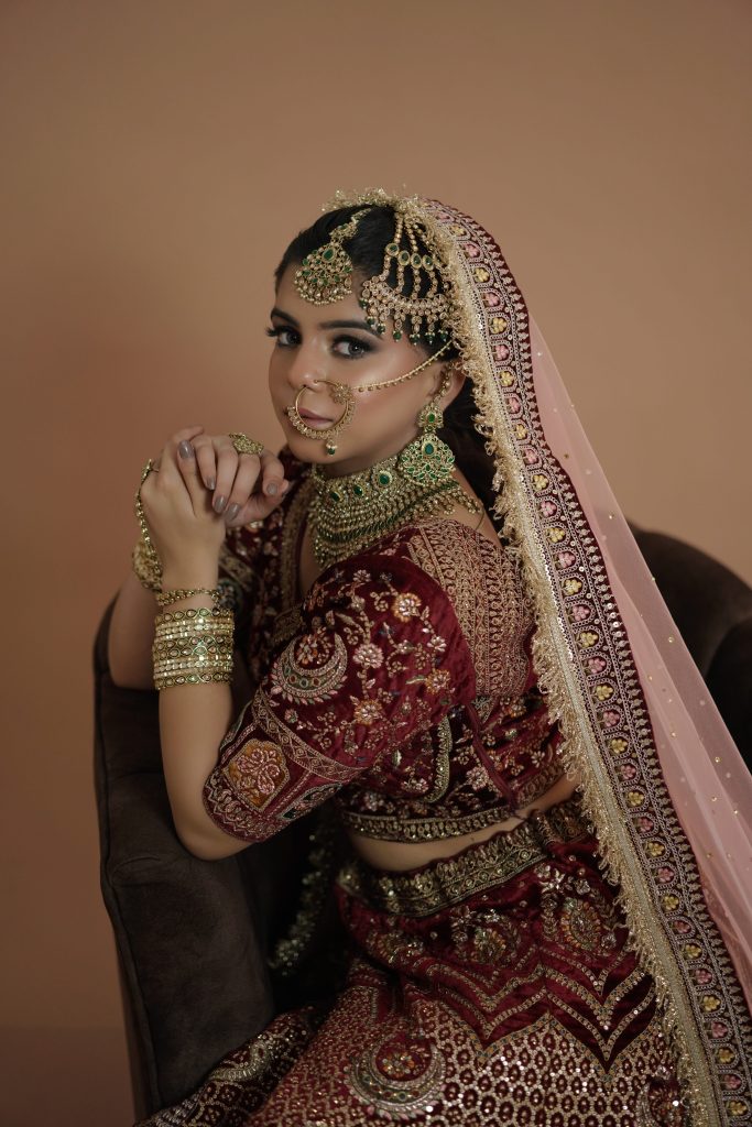 Bride wearing a maroon embroidered bridal lehenga with gold and green detailing and traditional bridal jewellery, showcasing bridal wear at All Eyes On U in Chandigarh and Mohali.
